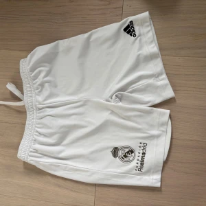 Vit Real Madrid kit  från Adidas - Säljer ett vitt Real Madrid set  från Adidas med klubbens logga och Adidas-märke. De har elastisk midja med snörning och är gjorda i mjuk polyester, perfekta för fotbollsträning eller match. Klassisk sportig look för dig som älskar fotboll.