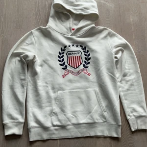 Vit GANT hoodie  - Vit hoodie från GANT med broderat emblem framtill i blått och rött. Klassisk känguruficka och huva. Mjuk insida och ribbade muddar. Perfekt för dig som gillar stilren och sportig look. Använd väldigt få antal gånger.