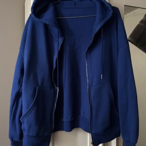 Blå hoodie med dragkedja och huva - Säljer en blå hoodie med dragkedja och huva. Tröjan har stora fickor framtill och ribbade muddar vid ärmslut och nederkant. Oversized passform som är perfekt för en chill och avslappnad stil. Materialet känns mjukt och skönt mot huden.