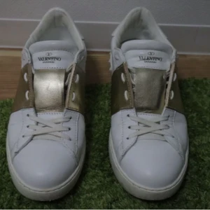 Valentino Garavani vita och guld sneakers - Snygga sneakers från Valentino Garavani i vitt skinn med breda guldfärgade detaljer över snörningen. Klassisk rund tå och platt sula. Perfekta för dig som vill sticka ut med en lyxig touch i vardagen.