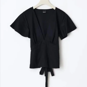 Svart omlottopp med knytband från gina - Svart omlottopp från Monki med djup v-ringning och korta, vida ärmar. Toppen har knytband i midjan för en snygg passform och är tillverkad i mjuk bomull. Perfekt för att skapa en trendig och avslappnad look.