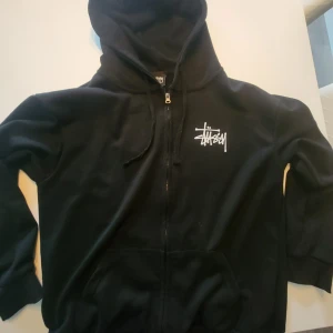 Svart Stüssy hoodie med dragkedja - Svart hoodie från Stüssy med vit logga på bröstet och stor logga på ryggen. Tröjan har huva med snörning, dragkedja framtill och känguruficka. Perfekt streetwear-plagg med skön passform och klassisk look. Den är så gott som ny. Storlek S. Priset är ej hugget i sten. Om du har mer frågor är det bara att skriva🤗. MVH Neo