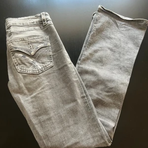 Grå utsvängda jeans med detaljerad bakficka från Gina - Snygga grå jeans med raka ben och coola broderade detaljer på bakfickan. Jeansen har klassisk femficksdesign och är perfekta för dig som gillar en stilren men ändå unik look. OBS knappar på bakfickorna har lossnat. Annars använda Max 3 gånger.