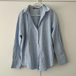 Blåvit randig skjorta från Zara - Säljer en oversized blåvit randig skjorta från Zara i bomull. Skjortan har klassisk krage, långa ärmar med breda manschetter och knappar framtill. Perfekt för en chill och stilren look. Pris 250, ordinarie 399kr