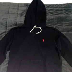 Svart hoodie från Ralph Lauren - Svart hoodie från Ralph Lauren med klassisk röd logga broderad på bröstet. Tröjan har huva med vita snören och en stor magficka. Perfekt för en avslappnad och stilren look.