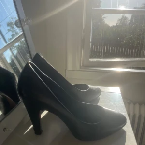 Svarta pumps med spetsig tå - Klassiska svarta pumps i skinn med spetsig tå och hög klack. Snygg och tidlös modell som passar till många olika outfits. Skorna har en elegant form och är tillverkade i ett slätt material som ger en stilren look.