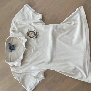 Beige  polo t-shirt från Ralph Lauren - Vit polo t-shirt från Ralph Lauren med klassisk krage och två knappar. Broderad logga och emblem i guld och marinblått på bröstet. Tillverkad i mjuk bomull, perfekt för en clean och stilren look.