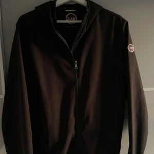 Svart vindjacka från Colmar - Svart vindjacka från Colmar med huva och dragkedja framtill. Jackan har en stilren look med Colmar-logga på ärmen och är tillverkad i ett slätt, lätt material. Perfekt för dig som vill ha en clean och sportig stil.