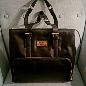 Svart datorväska från Skye & Soul - Stilren svart datorväska i skinn från Skye & Soul med guldfärgade metalldetaljer och dubbla handtag. Väskan har en stor ytterficka med dragkedja och rymligt huvudfack. Perfekt för att bära laptop och andra viktiga grejer.