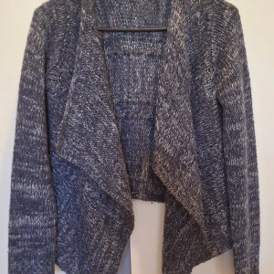 Grå stickad kofta från Lindex - Mysig gråmelerad stickad kofta från Lindex med öppen front och lång ärm. Koftan har en avslappnad passform och är tillverkad i ett garn med inslag av silvertråd som ger en diskret glittrig effekt. Perfekt för lager-på-lager under kyliga dagar. Funkar också för barn