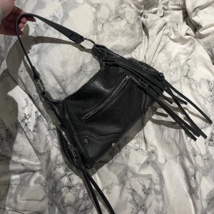 Svart axelväska med dragkedjor och fransar - Cool grå axelväska i skinn med flera dragkedjor och långa fransdetaljer. Väskan har både kort och lång rem, silverfärgade metalldetaljer den är andvänd en kväll så den är som ny!