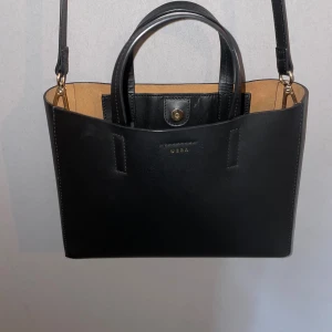 Svart toteväska i skinn från Wera - Stilren svart toteväska från Wera i äkta skinn. Väskan har både handtag och avtagbar axelrem i samma material, samt guldfärgade metalldetaljer. Insidan är ljusbrun och väskan stängs med tryckknapp. Väskan stängs med magnetknapp och kroklås i toppen. Inuti finns en enkel innerficka. PINZA-serien är stilren och elegant och finns i många fina modeller.  • Mindre toteväska i skinn • Stilren design • Smala topphandag • Avtagbar axelrem • En innerficka  Mått: Bredd: 27 cm Höjd: 19,5 cm Djup: 8 cm 