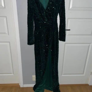 Grön glittrig långklänning med slits - Säljer en smaragdgrön långklänning med glittriga paljetter och v-ringning. Klänningen har långa ärmar, omlott-detalj framtill och en snygg slits som ger extra edge. Perfekt för dig som vill sticka ut på festen!