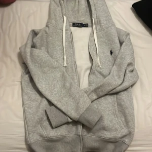 Grå hoodie från Polo Ralph Lauren - Snygg grå hoodie från Polo Ralph Lauren med dragkedja och vit snörning i huvan. Klassisk broderad logga på bröstet och mjukt material som känns riktigt skönt. Perfekt för dig som gillar en clean och stilren look.