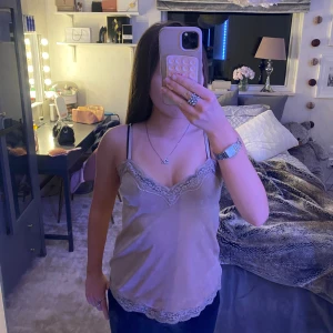 Beige linnetopp med spets från Hunkemöller - Supersöt beige linnetopp från Hunkemöller med tunna axelband och v-ringning. Toppen har fina spetsdetaljer längs halsringningen och nederkanten, och är gjord i ett glansigt, satinliknande material som känns lyxigt och mjukt mot huden.