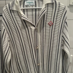 Randig stickad skjorta Casablanca - Säljer en stickad skjorta från Casablanca med vertikala svarta och grå ränder på vit botten. Skjortan har pärlknappar, krage och lång ärm. Märkets logga broderad på bröstet. Perfekt för dig som vill sticka ut med en unik och trendig look.