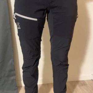 Svarta täckbyxor från Haglöfs - Svarta täckbyxor från Haglöfs med vita och svarta dragkedjor på benen. Byxorna har en smal passform och är tillverkade i slitstarkt syntetmaterial, perfekta för utomhusaktiviteter. Snygg logga på låret och flera praktiska fickor med dragkedja.