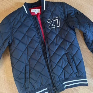 Quiltad bomberjacka Hampton Republic - Mörkblå quiltad bomberjacka från Hampton Republic med broderad siffra 27 på bröstet. Jackan har randiga muddar, dragkedja framtill och röd insida. Klassisk bombermodell med lång ärm och fickor på sidorna. Mycket bra skick, har endast blivit använd ett fåtal gånger.