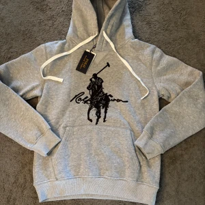 Grå hoodie från Polo Ralph Lauren - Snygg grå hoodie från Polo Ralph Lauren med klassiskt svart polospelartryck på bröstet. Tröjan har huva med vita snören, känguruficka och är tillverkad i 100% bomull för en skön och avslappnad känsla. Perfekt för chill dagar och streetstyle.