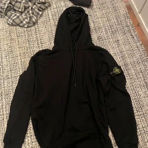Svart hoodie från Stone Island M - Svart hoodie från Stone Island med klassisk logga på vänster ärm. Tröjan har huva med snörning och en stor magficka. Tillverkad i mjuk bomull, perfekt för en chill och stilren look.