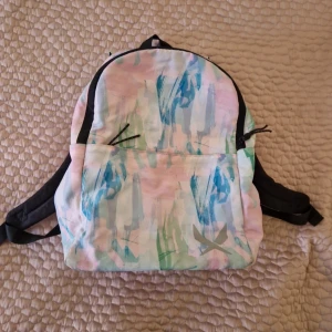 Pastellfärgad ryggsäck med mönster - Säljer en ryggsäck i pastellfärger med abstrakt mönster i blått, grönt och rosa. Väskan har svarta detaljer, justerbara axelband och ett stort huvudfack med innerficka. Materialet är troligtvis polyester och den har dragkedja upptill.