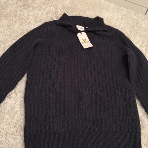 Mörkblå stickad tröja Lyle & Scott - Riktigt snygg mörkblå stickad tröja från Lyle & Scott med ribbad struktur och half zip-dragkedja vid kragen. Tröjan har lång ärm och en klassisk passform som funkar till många stilar. Perfekt för kyligare dagar.
