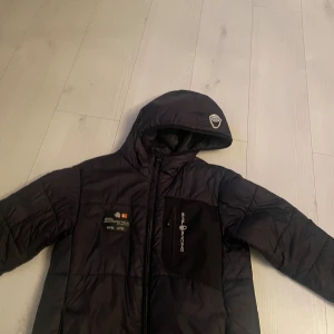 Svart pufferjacka från Sail Racing - Svart pufferjacka från Sail Racing med huva och dragkedja framtill. Jackan har en bröstficka med Sail Racing-logga och en snygg detalj på huvan. Tillverkad i syntetmaterial och perfekt för kyliga dagar.