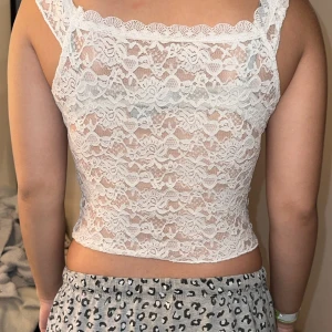 Vit spetsig croppad linnetopp - Säljer en vit croppad linnetopp i spets med v-ringning och liten rosett framtill. Toppen är ärmlös och har ett genombrutet spetsmönster som ger en romantisk vibe. Perfekt att styla med jeans eller kjol för en trendig look.