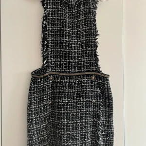 Svartvit rutig kort överallt med fransar - Säljer en svartvit kort klänning i rutigt mönster med råa fransade kanter och dekorativa knappar i midjan. Klänningen har en strukturerad känsla och är tillverkad i ett tjockare tyg som påminner om ullmix. Perfekt för dig som gillar en chic och edgy stil.