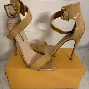 Beige Steve Madden klackskor - Snygga beige klackpumps från Steve Madden med hög klack och breda remmar över vrist och tår. Skorna har en stor guldfärgad spänne och är tillverkade i glansigt syntetmaterial. 
