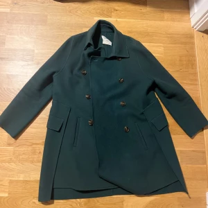 Mörkgrön skepparkappa från Oudifu - Stilren mörkgrön skepparkavaj från Oudifu med dubbelknäppning och klassisk krage. Jackan har två fickor med lock och är helfodrad. Perfekt för dig som vill ha en snygg och tidlös look under höst och vinter.