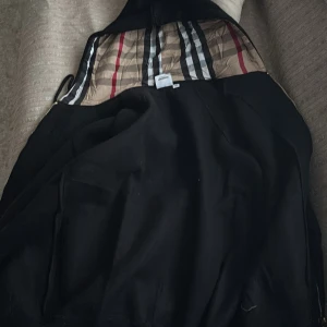 Svart vindjacka från Burberry - Svart vindjacka från Burberry med klassiskt rutigt foder i beige, rött och svart. Jackan har huva med snörning, dragkedja framtill och två fickor. Perfekt för dig som vill ha en stilren jacka med lyxig känsla och ikoniska Burberry-detaljer.