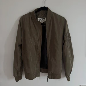 Olivgrön bomberjacka från Junkyard  - Snygg olivgrön bomberjacka från Junkyard i storlek XS. Jackan har klassisk ribbad krage, muddar och midja, samt dragkedja framtill och på ärmen. Tillverkad i polyester med en clean look och praktiska fickor. Perfekt för dig som gillar streetstyle.