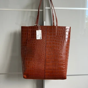 Brun croco shoppingväska från NA-KD - Stilren brun shoppingväska från NA-KD i croco-mönstrad syntet. Väskan har rejäla handtag, rymligt innerfack och en mindre ficka på insidan. 