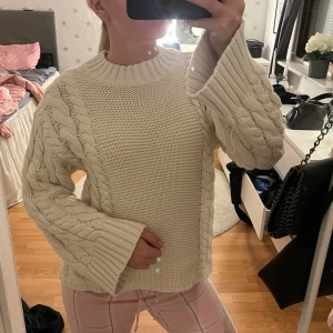 Beige stickad tröja med flätmönster - Mysig beige stickad tröja med breda ärmar och snyggt flätmönster längs sidorna och ärmarna. Tröjan har en grovstickad struktur och en hög ribbad krage.