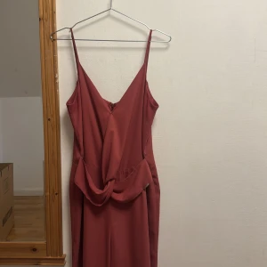 Vinröd långklänning med knutdetalj - Säljer en vinröd långklänning med smala axelband och snygg v-ringning. Klänningen har en elegant knutdetalj i midjan och dragkedja bak. Materialet är slätt och faller fint längs kroppen, perfekt för dig som vill ha en stilren och trendig look.