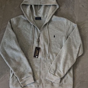 Grå hoodie från Polo Ralph Lauren - Snygg grå hoodie från Polo Ralph Lauren med dragkedja och huva. Klassisk logga broderad på bröstet och praktiska fickor framtill. Tillverkad i mjuk bomullsmix, perfekt för chill dagar. Stilsäker och enkel att matcha med allt.