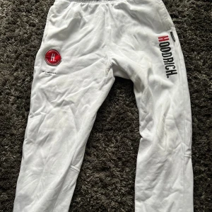 Vita mjukisbyxor från Hoodrich - Snygga vita mjukisbyxor från Hoodrich med tryckt logga i svart och rött längs benet samt ett broderat märke på höften. Byxorna har elastisk midja och är gjorda i mjukt bomullsmaterial, perfekta för chill eller streetwear.