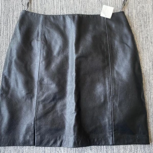 Svart skinnkjol med slits - Snygg svart kort skinnkjol med slitsar fram och diskret dragkedja bak. Kjolen har en stilren design med slät yta och raka linjer, perfekt för dig som gillar minimalistisk stil. Materialet är mjukt och följsamt, vilket gör den bekväm att bära. Mått Midjan: ca 39 cm Total längd: ca 47 cm  100% läder, kjol och 100% polyester, underkjol  Kommer från ett djur och rökfritt hem.