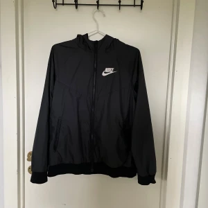 Svart vindjacka från Nike  - Svart vindjacka från Nike i storlek Xs med vit logga på bröstet. Jackan har huva, hel dragkedja framtill och två fickor med dragkedja. Tillverkad i lätt polyester, perfekt för blåsiga dagar. Sportig och stilren design.