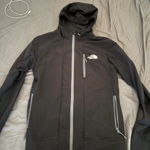 Svart vindjacka från The North Face - Svart vindjacka från The North Face med huva och tre grå dragkedjor framtill. Jackan har logga på bröstet och är långärmad med en clean, sportig look. Perfekt för blåsiga dagar och outdoor-äventyr.
