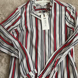 Randig skjorta från Holly & Whyte - Snygg skjorta med vertikala ränder i vitt, svart och rött från Holly & Whyte. Modellen har lång ärm med knappar vid manschetten och klassisk krage. Skjortan är gjord i mjuk bomull och har en relaxed passform som är enkel att styla till jeans eller kjol.