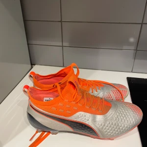 Puma fotbollsskor i orange och silver - Säljer ett par grymma Puma fotbollsskor i en riktigt snygg kombo av orange och silver. Skorna har snörning, mönstrad ovandel och klassisk svart Puma-logga på sidan. Perfekta dobbar för spel på gräs och en tajt passform som ger bra bollkontroll.