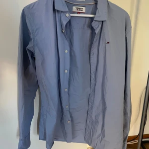 Blå skjorta från Tommy Jeans - Snygg ljusblå skjorta från Tommy Jeans i slim fit-modell. Skjortan har klassisk krage, knäppning framtill och diskret logga på bröstet. Tillverkad i bomull och med långa ärmar
