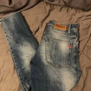 Blå slitna Diesel jeans, storlek 30 - Säljer ett par blå jeans från Diesel med snyggt slitna detaljer och en liten reva på framsidan. Klassisk femficksmodell med läderpatch bak och cool tvättad look. Passar dig som gillar street och avslappnad stil.
