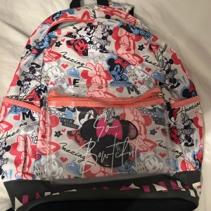 Disney Minnie Mouse ryggsäck färgglad - Färgglad ryggsäck med Minnie Mouse-tryck i rött, blått, svart och vitt. Väskan har ett stort huvudfack, framficka i transparent plast med texten 'So Bowtiful' och orange dragkedjor. Justerbara axelband och vadderad baksida för extra komfort.