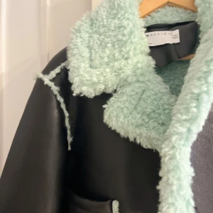 Svart och mintgrön teddyjacka från asos-design - Säljer en svart croppad teddyjacka från asos-design med mintgrönt fuskpälsfoder och detaljer. Jackan har stora fickor, bred krage och långa ärmar med mjuka teddykanter. Perfekt statement-plagg för dig som vill sticka ut.