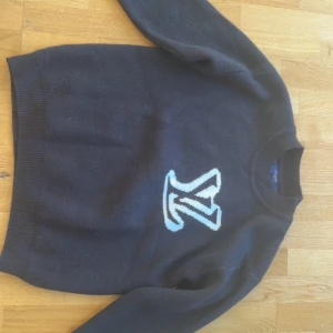 Svart Louis Vuitton stickad tröja - Snygg svart stickad tröja från Louis Vuitton med stort LV-logo i vitt och blått framtill. Tröjan har rund halsringning och långa ärmar, perfekt för dig som vill ha en klassisk men ändå trendig look.