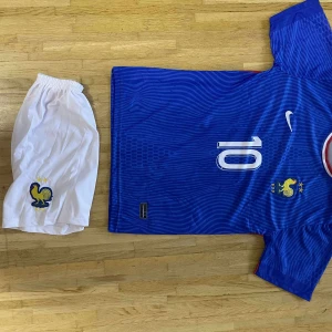 Frankrike Mbappé fotbollströja Nike - Säljer en blå fransk landslagströja från Nike med Mbappé och nummer 10 på ryggen. Tröjan har vita detaljer, röda inslag på axlarna och gult tuppemblem på bröstet. Matchande vita shorts med tuppemblem ingår. Tillverkad i lätt och ventilerande material.