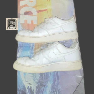 Nike Air Force 1 vita sneakers - Klassiska Nike Air Force 1 sneakers i helvitt skinn med rund tå och platt sula. Modellen har snörning och den ikoniska swoosh-loggan på sidan. Perfekt för dig som gillar clean och tidlös streetstil.